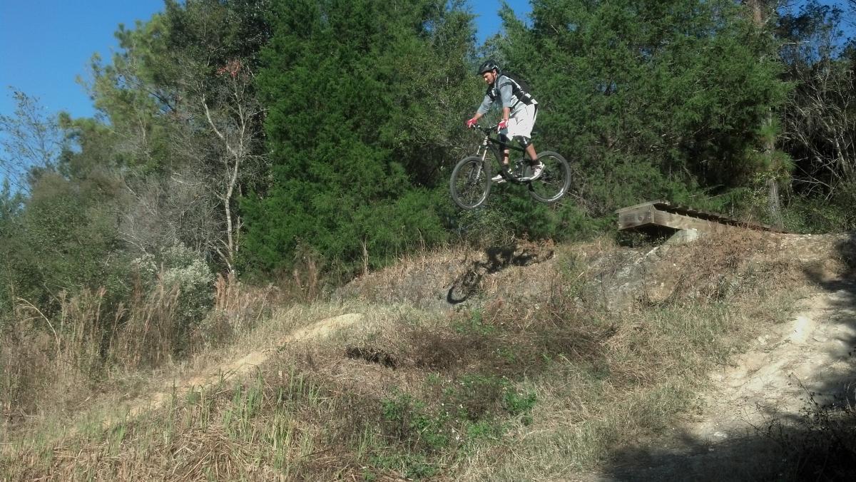 A mountain biker in mid-air, jumping over a dirt ramp surrounded by trees and shrubs. The rider is wearing a helmet, gloves, and a light-colored outfit, showcasing an action-packed moment in a natural outdoor setting. Santos mountain bike trail.