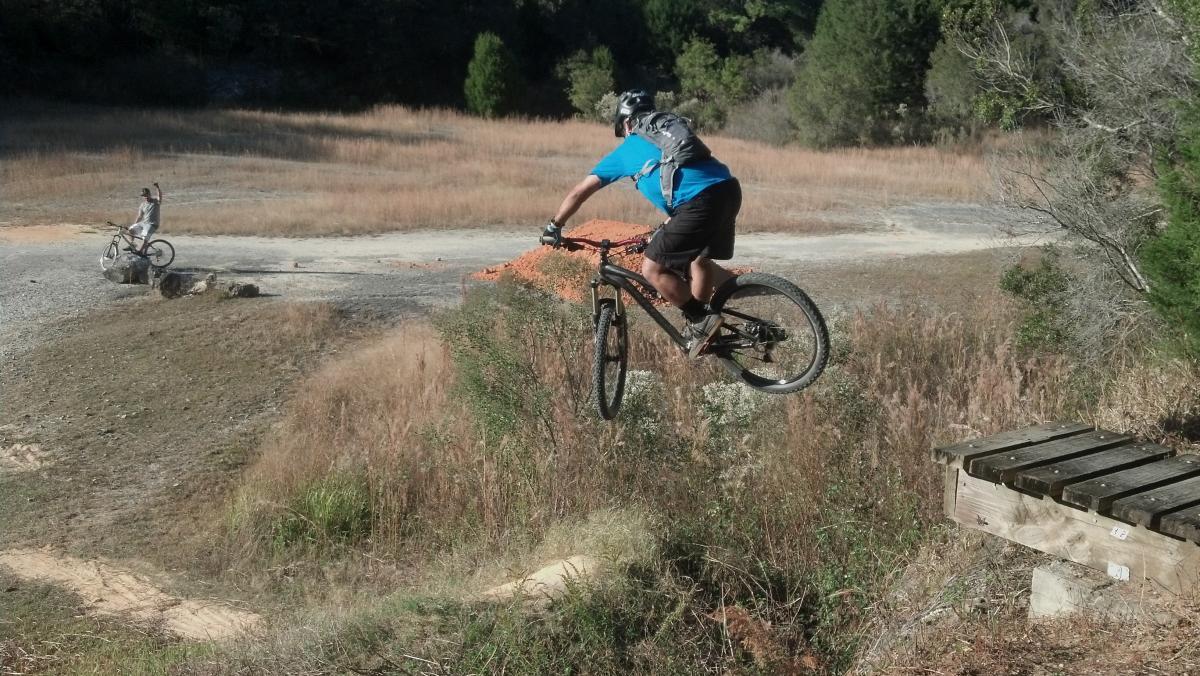 A mountain biker in a blue shirt and black shorts performs a jump over a small wooden ramp while riding on an uneven, grassy terrain. In the background, another cyclist is seen navigating the trail. The scene is surrounded by sparse vegetation under a clear sky. Santos mountain bike trail.