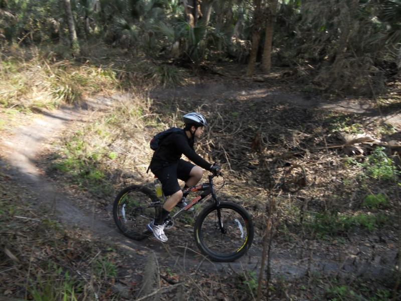 A cyclist in a black outfit and helmet rides a mountain bike along a narrow dirt trail in a wooded area with sparse vegetation and trees. Chuck Lennon Park mountain bike trail.