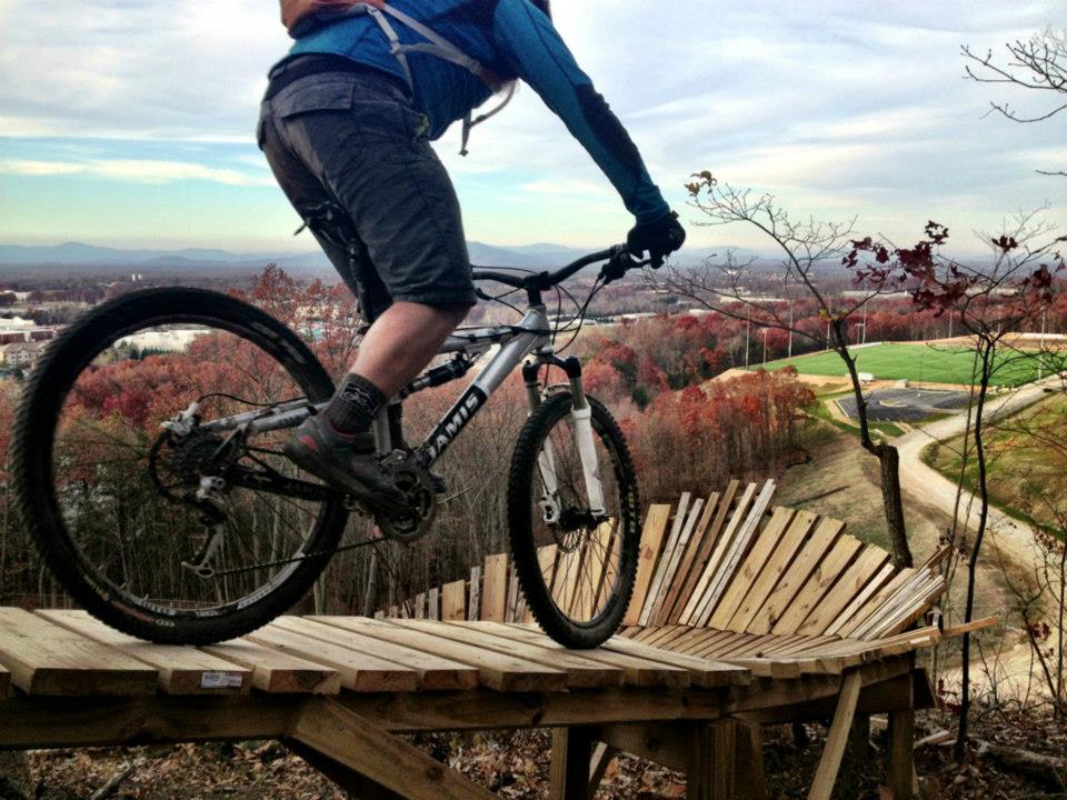 Jamis Dakar XLT: A mountain biker riding a wooden ramp on a hillside, surrounded by autumn-colored trees and a scenic view of a valley below.