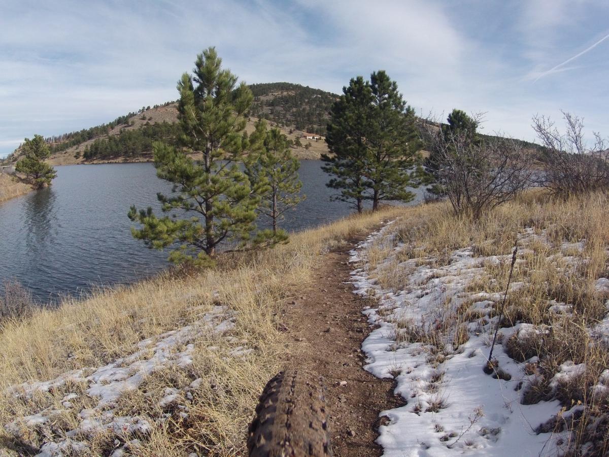 A scenic view of a lake surrounded by a mountainous landscape, with a dirt path leading along the water's edge. Pine trees frame the scene, and patches of snow are visible along the grassy trail, suggesting a cool, fresh atmosphere. The sky is partially cloudy, enhancing the natural beauty of the environment. Pinewood Reservoir mountain bike trail.