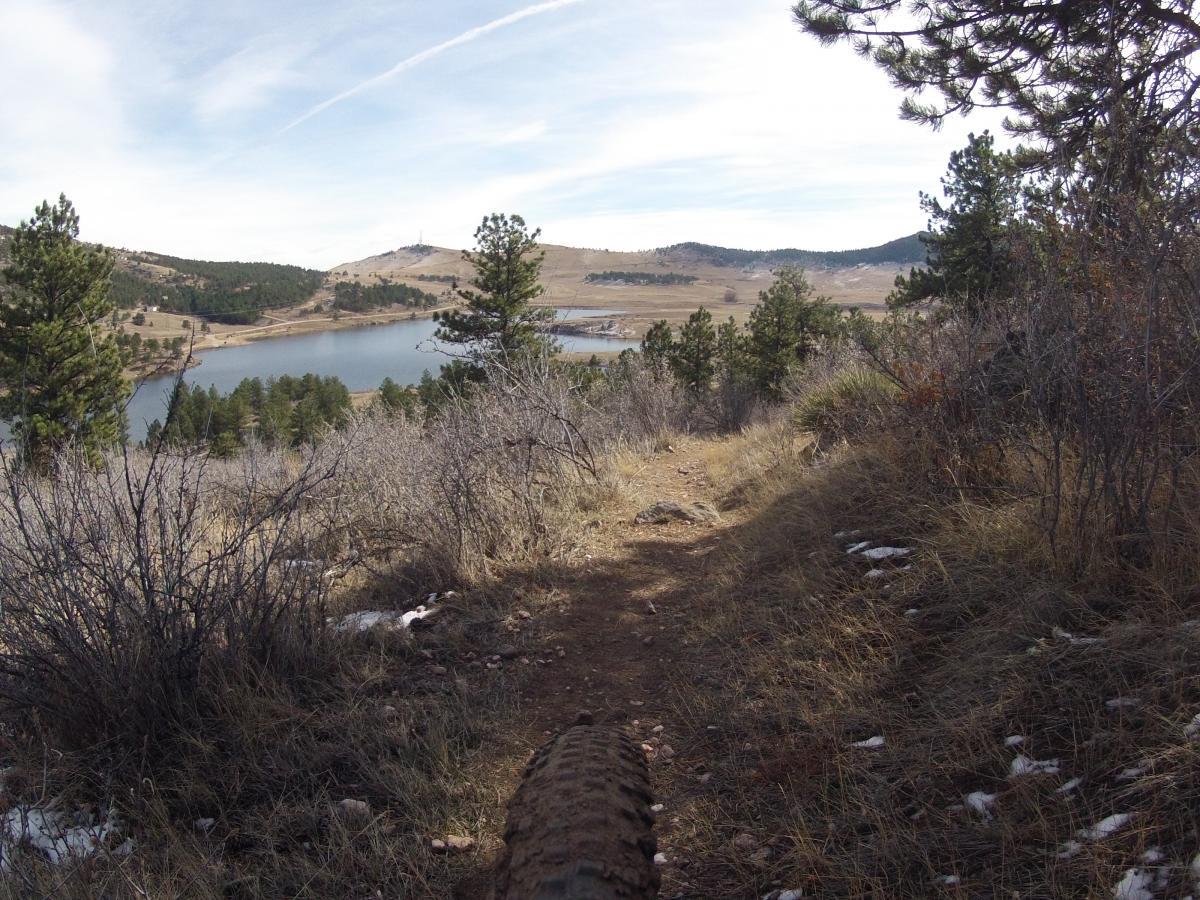 A scenic view of a winding dirt trail surrounded by tall trees, overlooking a calm lake and rolling hills in the background. The landscape features green pine trees and sparse underbrush, with patches of snow visible on the ground. The sky is partly cloudy with trails from aircraft. Pinewood Reservoir mountain bike trail.