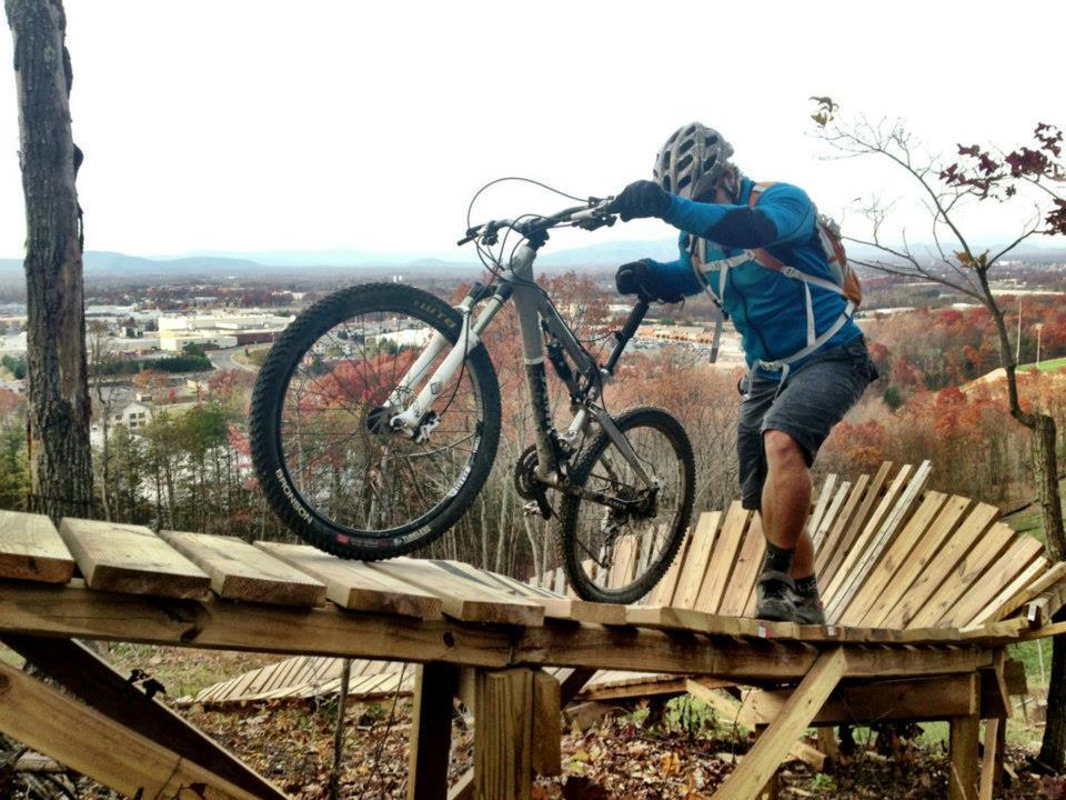 Jamis Dakar XLT: A mountain biker skillfully navigates a wooden rollercoaster-style trail, lifting the front wheel of his bike off the ground. He is wearing a blue long-sleeve shirt, a helmet, and a backpack, with a scenic view of colorful autumn foliage and a town in the background.