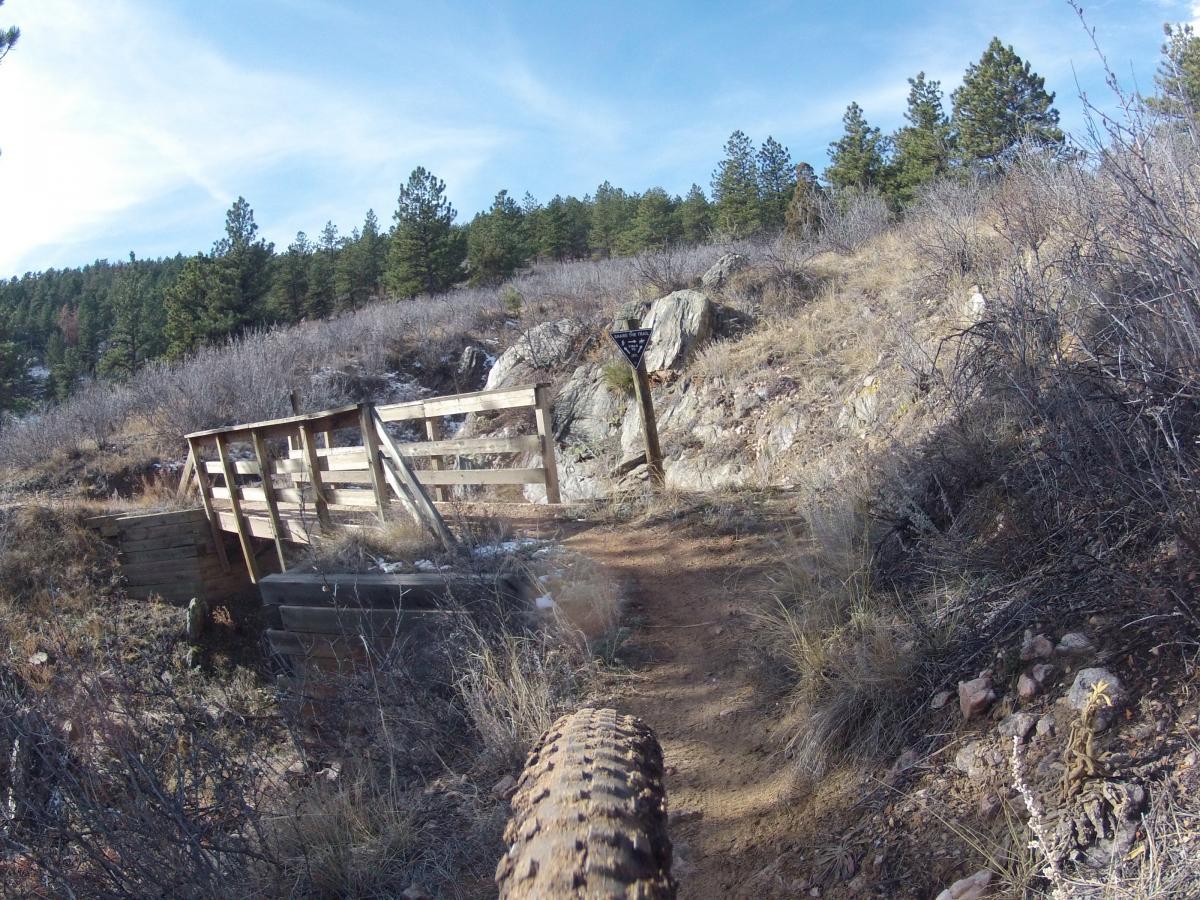 A view of a winding dirt trail leading to a wooden bridge, surrounded by sparse vegetation and rocky terrain under a blue sky. A trail sign is visible in the background, indicating direction or information about the path. The image captures the perspective from a mountain bike tire on the trail. Pinewood Reservoir mountain bike trail.