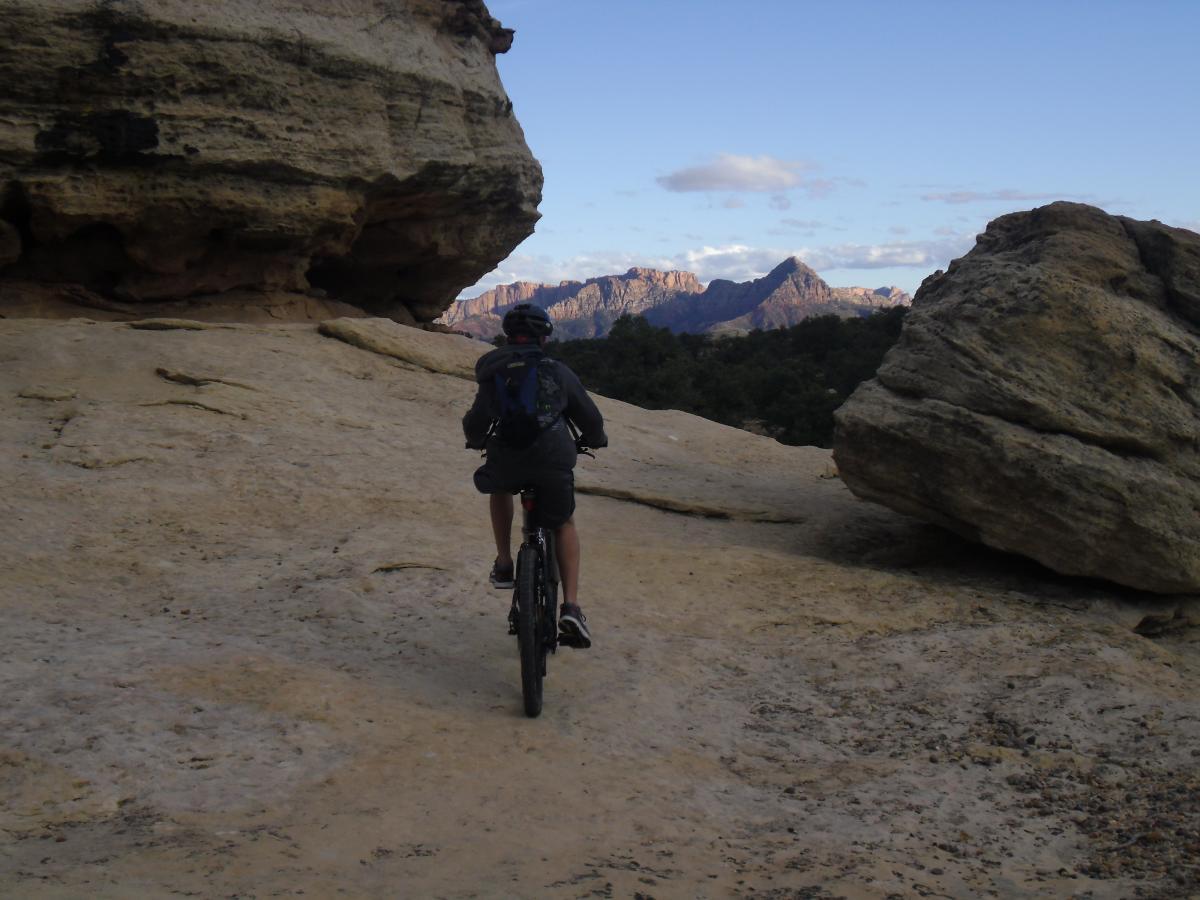 A person riding a mountain bike on a rocky trail, surrounded by large boulders and distant mountains under a clear sky. Gooseberry Mesa mountain bike trail.