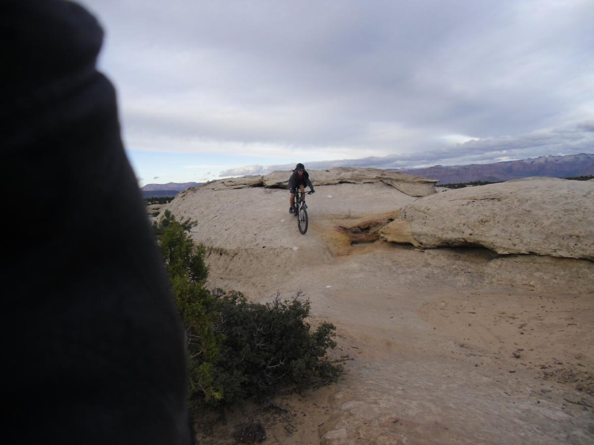 A mountain biker navigating a rocky terrain on a cloudy day, with a panoramic view of mountains in the background. The biker is riding along a plateau, showcasing a combination of skill and adventure amidst the natural landscape. Gooseberry Mesa mountain bike trail.