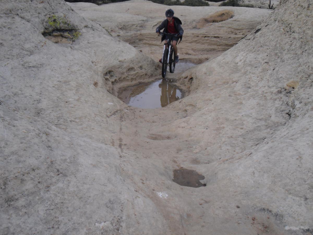 A person riding a mountain bike along a rocky trail with uneven terrain and a small puddle of water visible. The surroundings feature smooth, light-colored rock and sparse vegetation in the background. Gooseberry Mesa mountain bike trail.