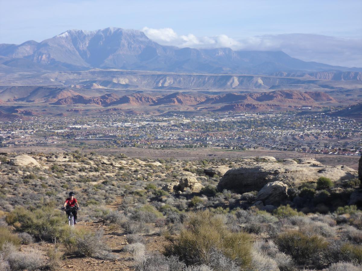A cyclist rides along a rugged trail in a desert landscape, with shrubs and rocky terrain in the foreground. In the background, expansive mountain ranges and a small town are visible beneath a partly cloudy sky. Zen Trail mountain bike trail.