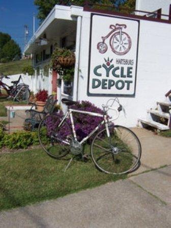 A white bicycle leaning against a wall with a sign that reads "Harrisonburg Cycle Depot." The building is surrounded by colorful plants and flowers, with additional bikes visible nearby. The scene is set on a sunny day.