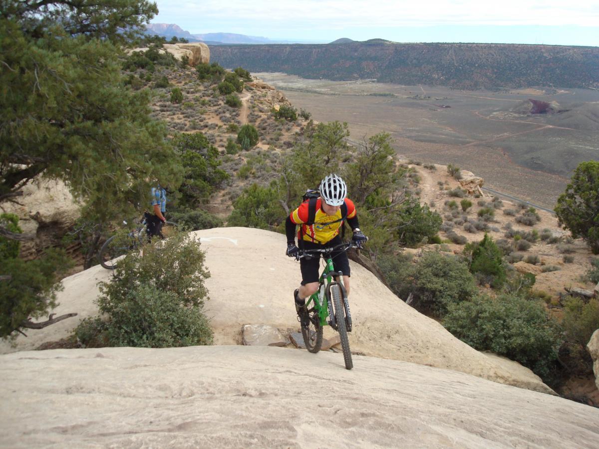 A mountain biker ascending a rocky incline in a desert landscape, surrounded by shrubs and trees, with another cyclist visible in the background. The scene showcases a rugged trail with distant mountains under a cloudy sky. Gooseberry Mesa mountain bike trail.