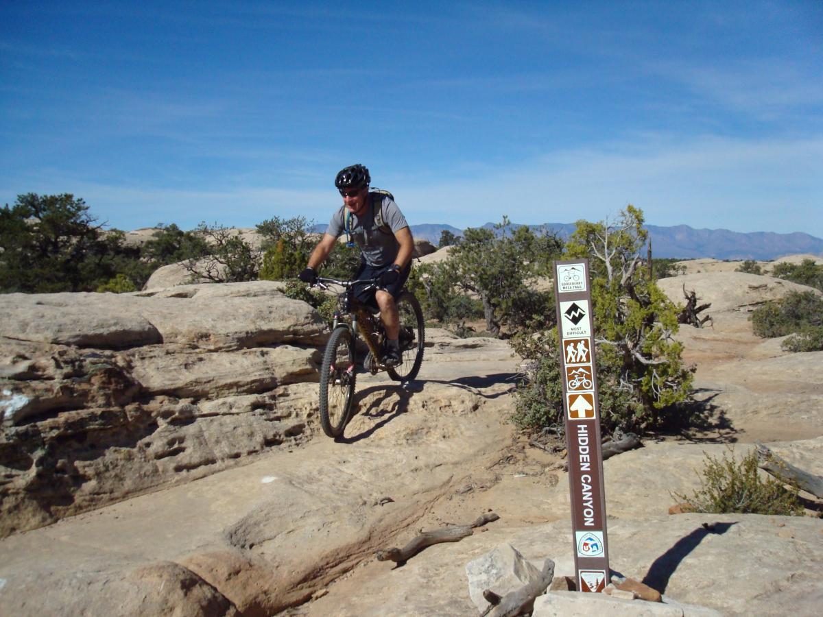 A mountain biker navigating a rocky trail in a desert landscape, with a signpost indicating the direction to "Hidden Canyon." The background features a clear blue sky and scattered shrubs. Gooseberry Mesa mountain bike trail.