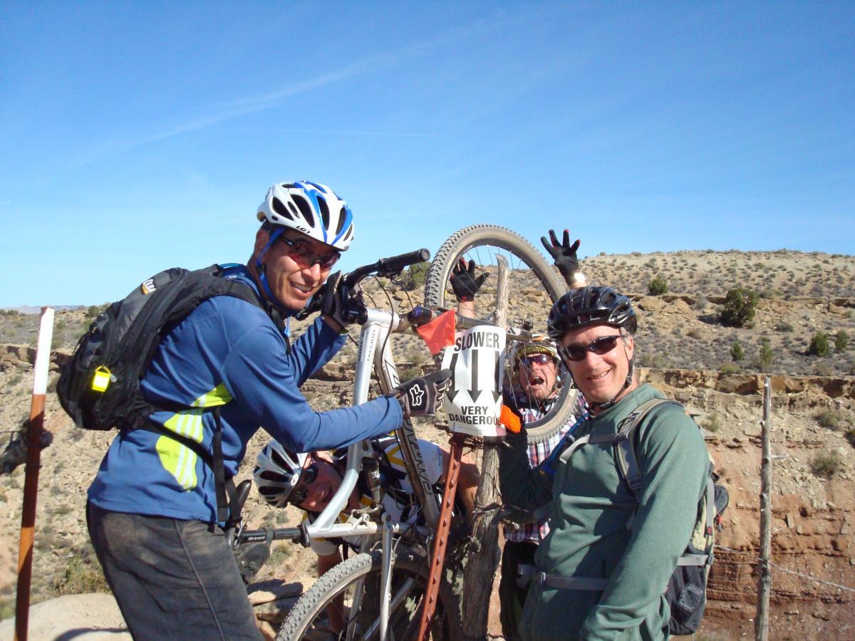 Two men wearing cycling gear smile for the camera while posing with a mountain bike. One man holds the bike up and points to a sign that reads "SLOWER, VERY DANGEROUS." In the background, a third person playfully peeks out from behind the bike. The scene is outdoors with a rocky landscape and clear blue sky. J.E.M. Trail mountain bike trail.
