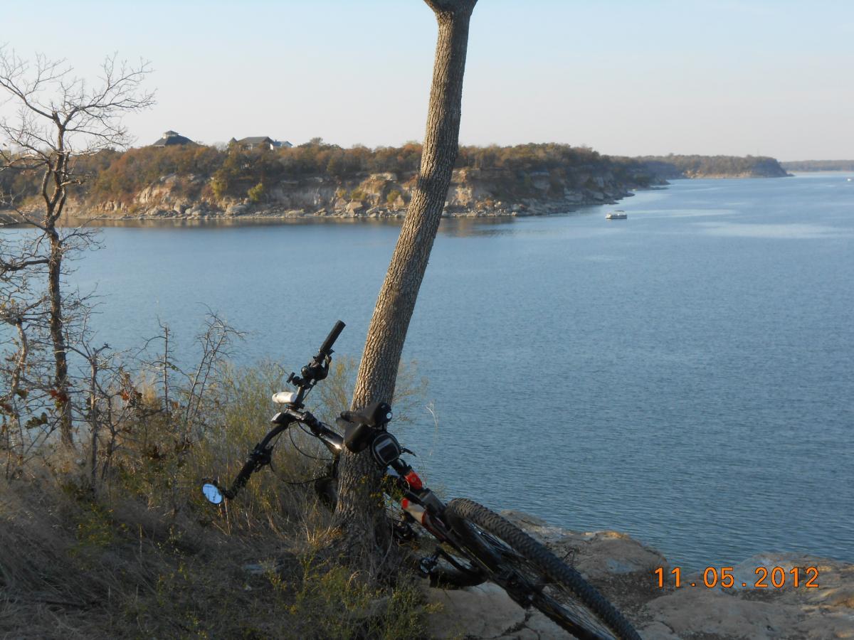 A mountain bike leaning against a tree, with a scenic view of a lake and rocky cliffs in the background. The scene is tranquil, depicting the natural landscape during the late afternoon or early evening, with clear skies and calm water. Eisenhower State Park mountain bike trail.