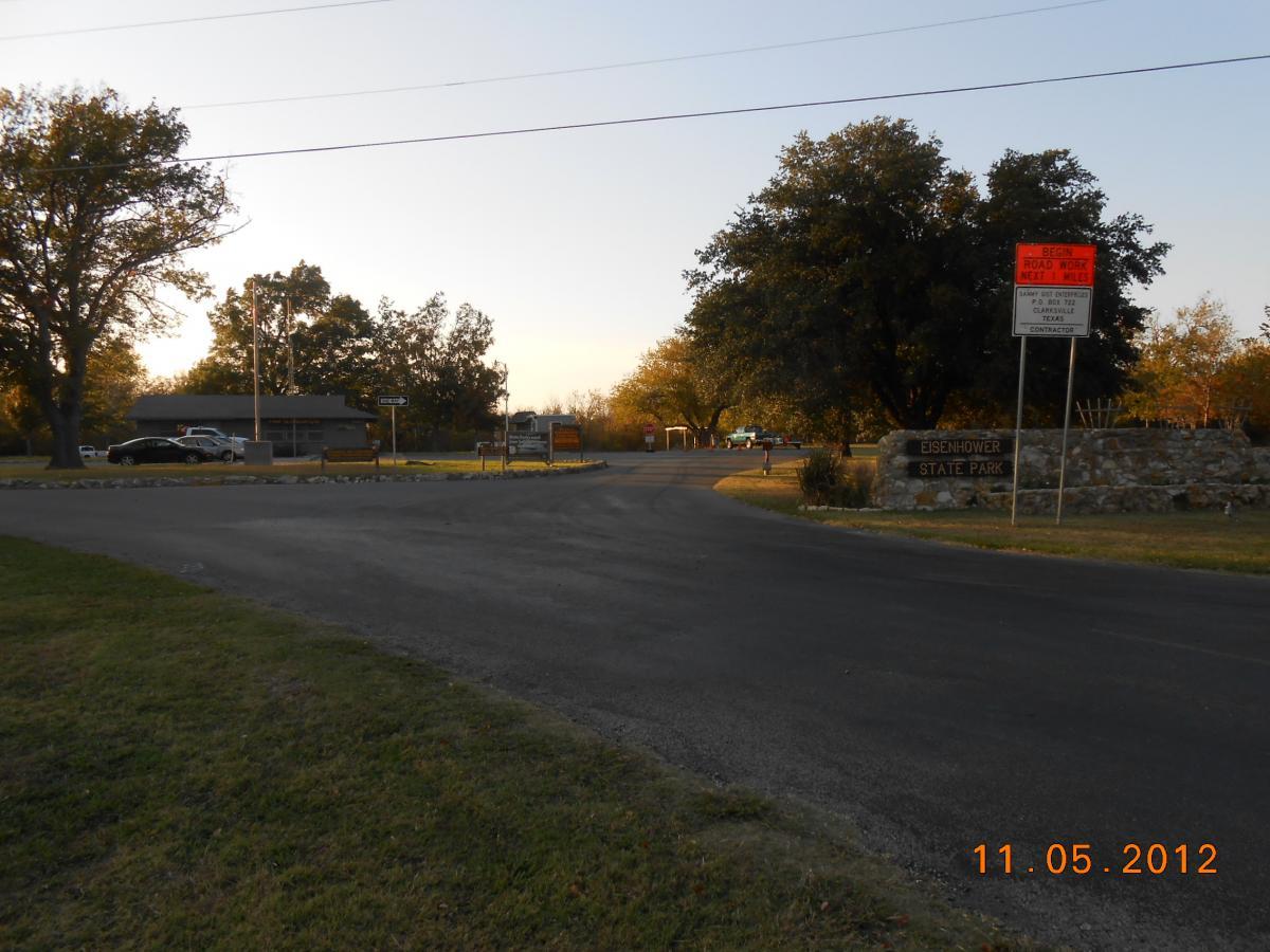 A view of the entrance to Eisenhower State Park, featuring a curved road leading into the park. There are trees on either side, a parking area with vehicles, and a stone sign indicating the park's name. A road work sign is visible, warning of upcoming road work nearby. The image is taken during golden hour, with soft lighting illuminating the scene. Eisenhower State Park mountain bike trail.