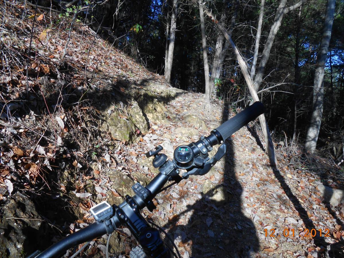 Alt text: Close-up view of a mountain bike handlebar on a dirt trail surrounded by fallen leaves and trees. The photo captures the bike's gear shifters and a small compass, with sunlight filtering through the forest. The Ridgeland Trails mountain bike trail.