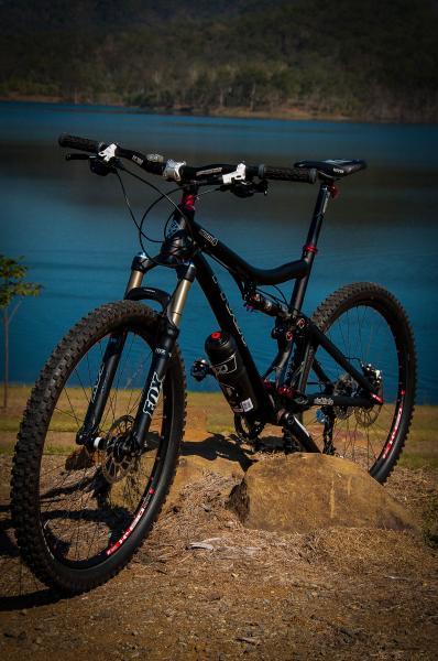 Pivot Mach 4: A mountain bike resting on a rock near a tranquil lake, surrounded by greenery. The bike features thick tires, front suspension forks, and a water bottle holder, with the water reflecting the surrounding landscape.
