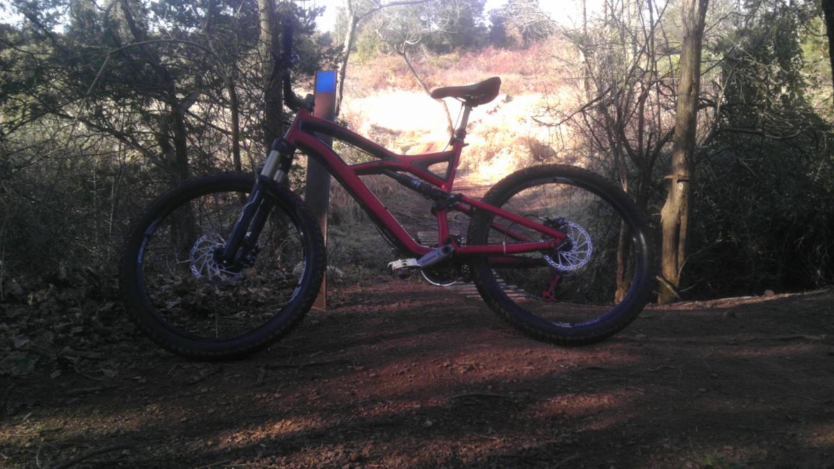 A red mountain bike is positioned on a dirt trail surrounded by trees and underbrush. The bike is angled to showcase its frame and wheels, with sunlight filtering through the foliage in the background. Six Mile Run mountain bike trail.