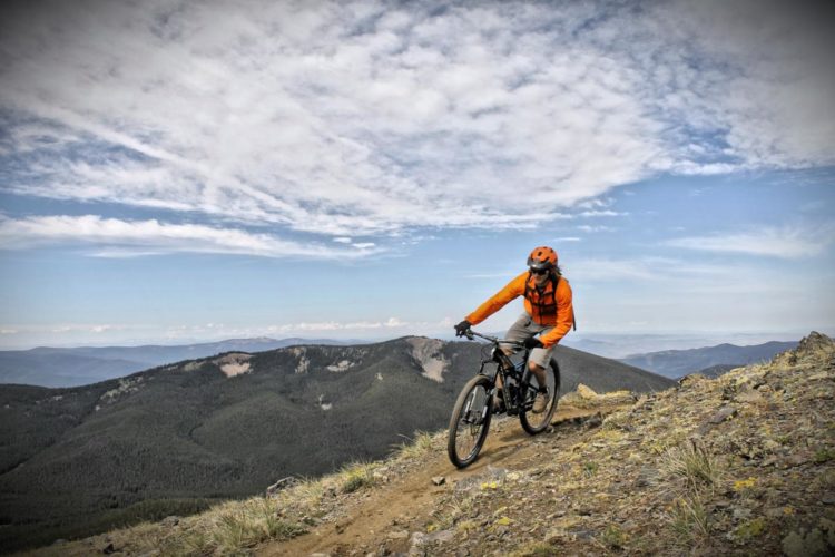 A mountain biker wearing an orange jacket and helmet rides along a narrow trail with a scenic view of hills and mountains in the background under a partly cloudy sky.
