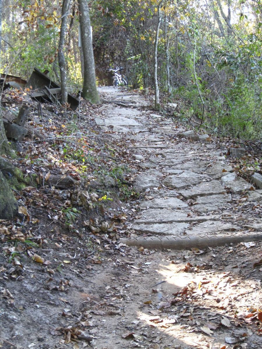 A winding, rocky path through a wooded area, surrounded by trees and scattered autumn leaves. A bicycle can be seen parked in the background, partially obscured by vegetation. The scene captures a tranquil and natural setting. Cadillac mountain bike trail.
