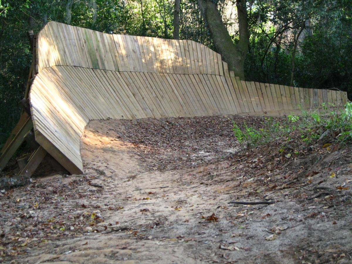 A wooden bike ramp or pump track featuring a curved design, surrounded by a natural forest setting with trees and scattered leaves on the ground. The ramp has a smooth surface made of wooden planks and is built on a dirt path. Tom Brown / Lafayette Heritage Park mountain bike trail.