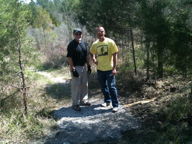 Two men standing on a gravel path in a wooded area. One man is wearing a black shirt and gray pants, while the other is dressed in a yellow shirt and jeans. They are smiling and looking at the camera, surrounded by greenery and trees. Butts Park mountain bike trail.