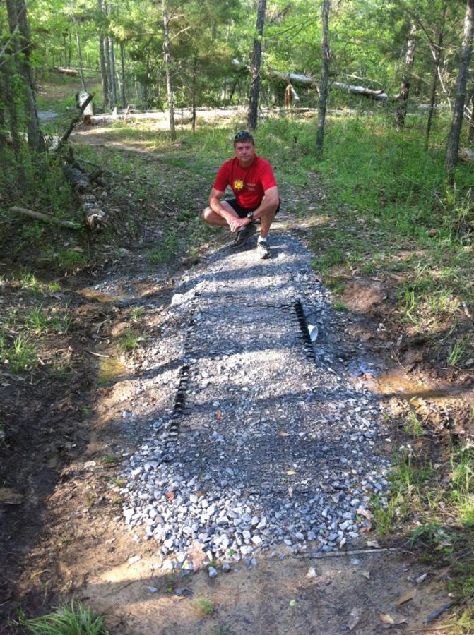 A man squatting on a newly constructed gravel pathway that crosses a muddy area in a wooded setting. Trees and greenery surround the path, indicating a natural outdoor environment. Butts Park mountain bike trail.