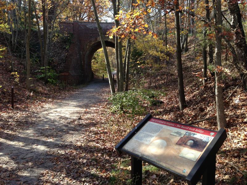 A scenic trail winding through autumn foliage, leading to a brick arched bridge in the background. In the foreground, a descriptive informational sign stands beside the path, detailing the history of the bridge. The ground is covered with fallen leaves, and trees line the sides of the trail, creating a tranquil outdoor setting. Laurel Hill Park mountain bike trail.