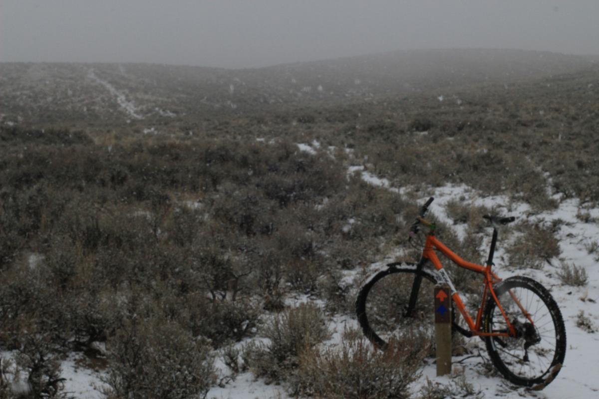An orange mountain bike is leaning against a signpost in a snowy, desolate landscape. The ground is covered with patches of snow, and sparse vegetation is visible in the foreground. In the background, rolling hills are partially obscured by a foggy, gray sky. Snowflakes are falling gently, creating a cold and quiet atmosphere. Discovery Hill Trails mountain bike trail.