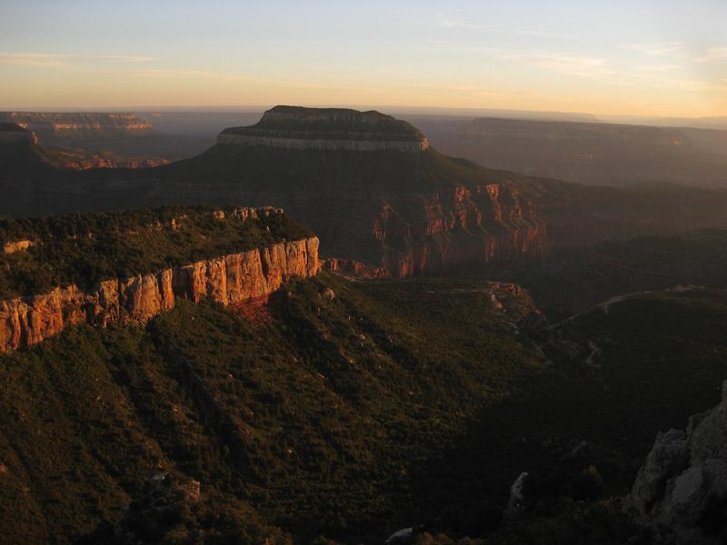 A scenic view of a rocky landscape at sunset, featuring layered cliffs and a prominent plateau surrounded by deep canyons. The warm colors of the setting sun illuminate the rugged terrain, highlighting the natural beauty of the area. Rainbow Rim Trail mountain bike trail.