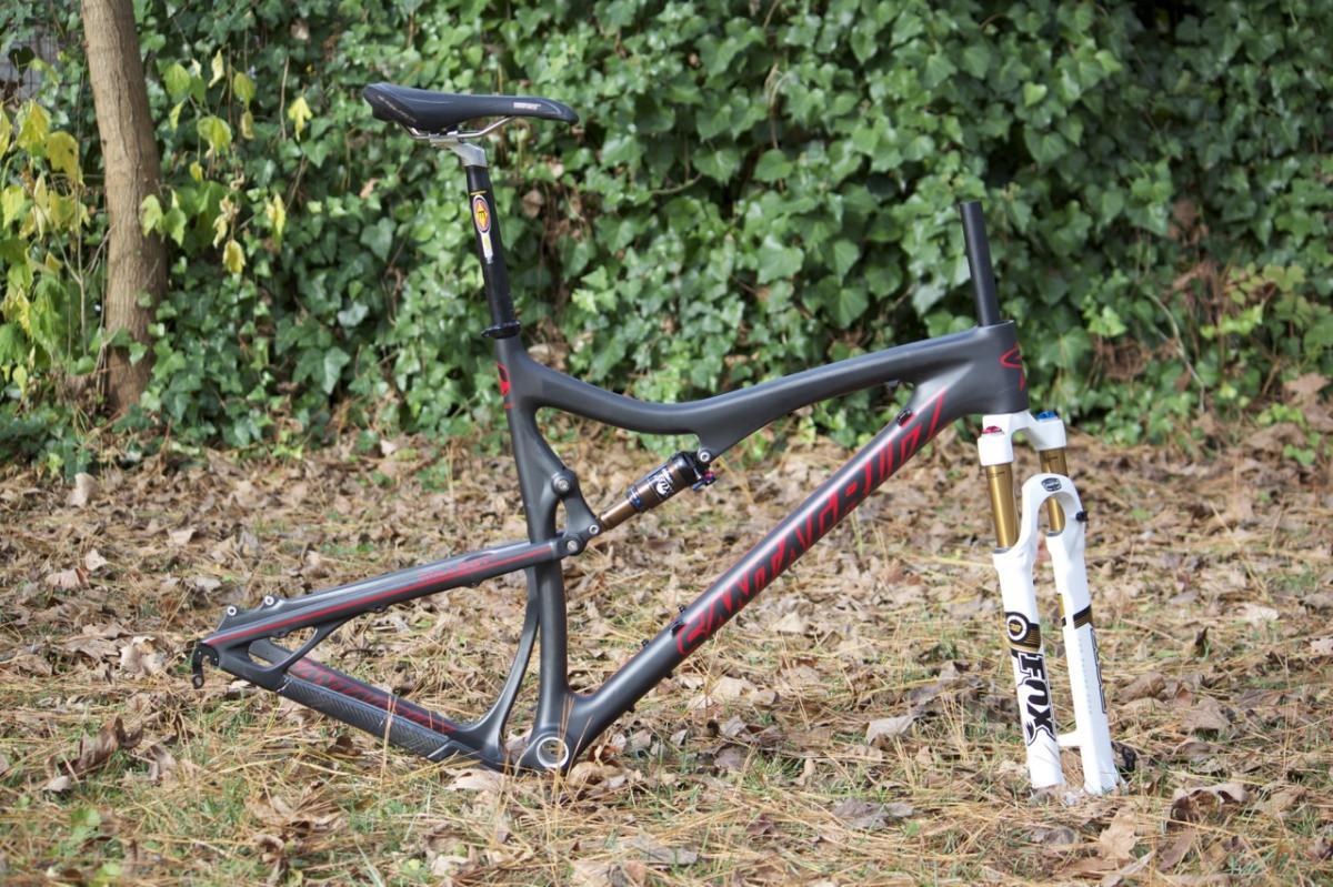 Santa Cruz Tallboy Carbon: A bicycle frame in a natural outdoor setting, resting on a bed of fallen leaves. The frame is primarily black with red accents and features a suspended rear shock. Attached to the front is a white and gold bicycle fork, indicating it is designed for performance. Lush green foliage provides a backdrop.
