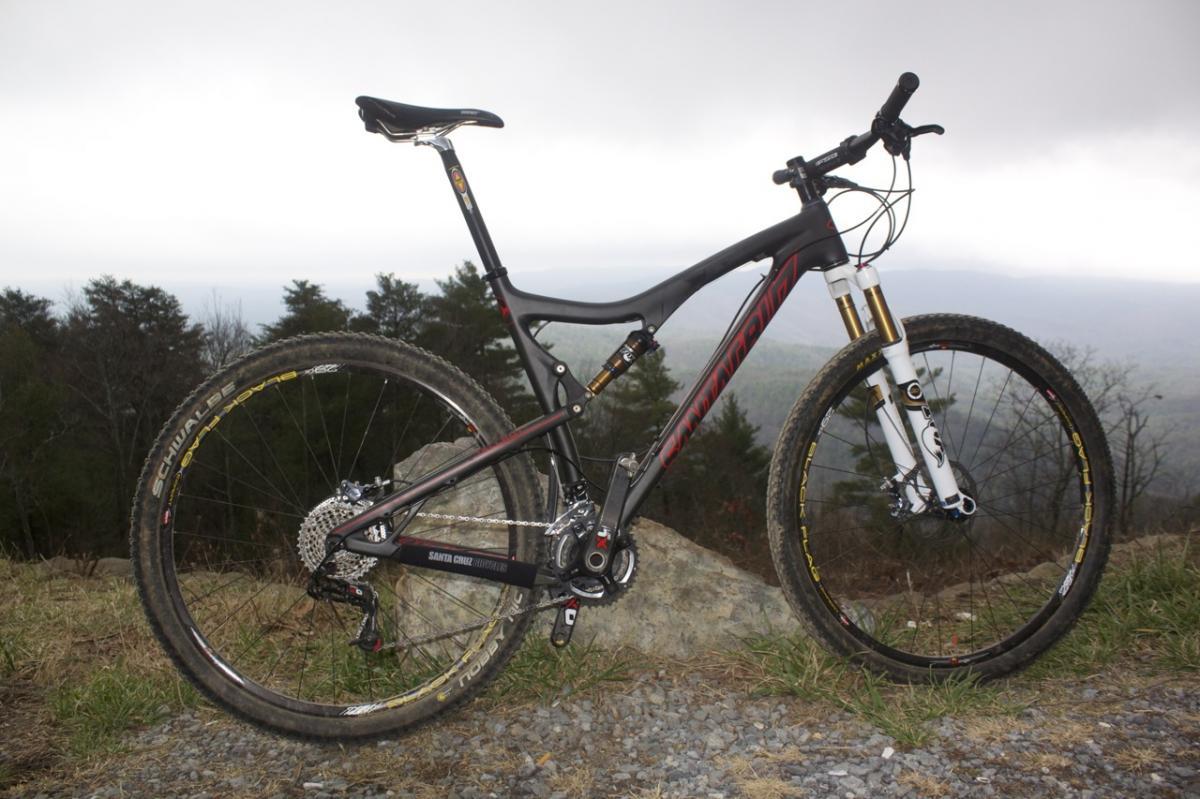 Santa Cruz Tallboy Carbon: A mountain bike positioned on a rock with a scenic view in the background, featuring a sleek black and red frame, front suspension fork, and knobby tires. The sky is overcast, suggesting a cool, outdoor riding environment.