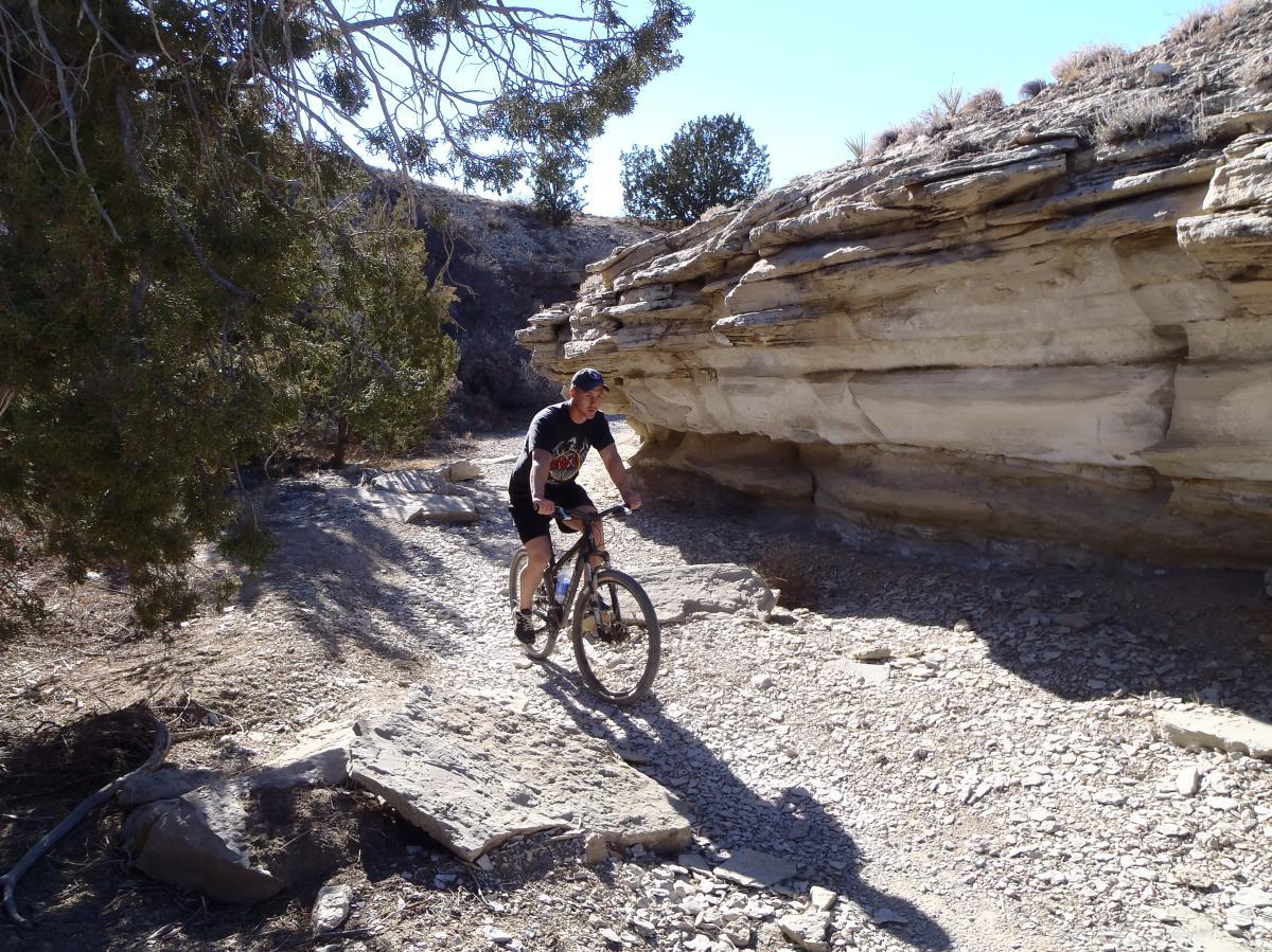 A mountain biker riding along a rocky trail surrounded by steep cliffs and sparse vegetation under a clear blue sky. South Shore Lake Pueblo mountain bike trail.