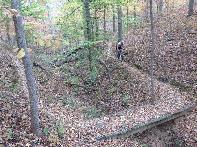 A mountain biker navigating a wooden bridge over a ravine in a wooded area, surrounded by trees with autumn foliage and leaves covering the ground. Modoc (Stevens Creek Trail) mountain bike trail.