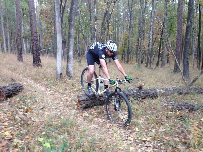 A cyclist in a black outfit navigates a challenging trail by riding over a fallen log in a wooded area. The background features tall trees with autumn foliage and a grassy path. Modoc (Stevens Creek Trail) mountain bike trail.
