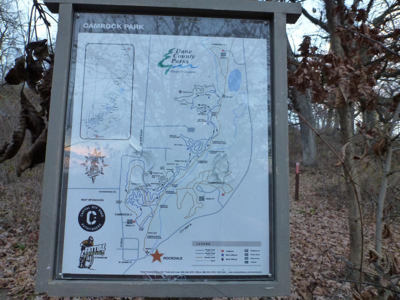 Map of Camrock Park in Dane County, showing trails, features, and point of interest locations within the park. The map includes a legend and highlights the Rockdale area. Surrounding the map are trees and fallen leaves, indicating a natural setting. Camrock 3 mountain bike trail.