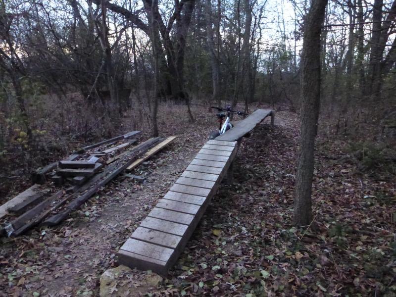 A narrow wooden bike ramp extending through a wooded area, with fallen leaves scattered on the ground. On one side, there are piles of assorted wood planks. A bicycle is resting against the ramp. The scene is dimly lit, suggesting early evening or dusk. Camrock 3 mountain bike trail.