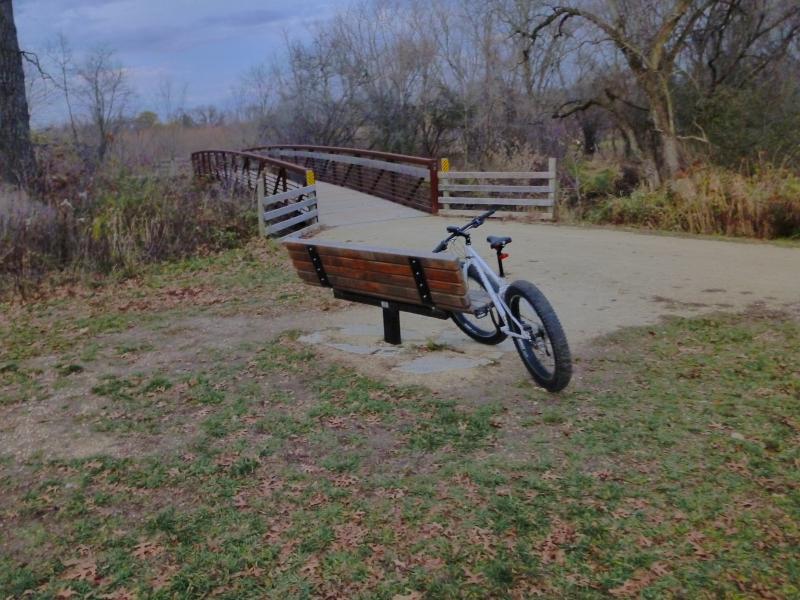 A mountain bike resting against a wooden bench in a park, with a walking bridge and grassy path visible in the background. The scene is surrounded by trees and autumn foliage. Camrock 3 mountain bike trail.
