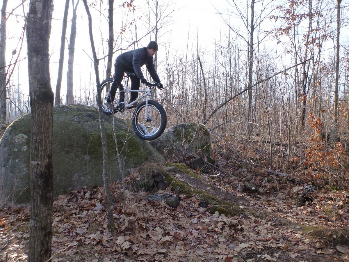 A person riding a fat tire mountain bike, skillfully balancing on top of a large rock in a wooded area during autumn. The ground is covered with fallen leaves, and the trees are bare, suggesting a cool season. Nicolet Nat'l Forest mountain bike trail.