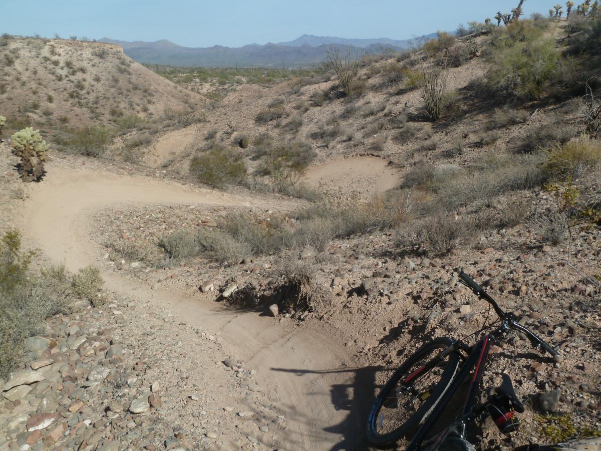 A winding dirt bike trail surrounded by sparse vegetation and rocky terrain, with a mountain backdrop under a clear blue sky. A mountain bike is resting on the ground in the foreground, indicating a biking adventure in a desert-like landscape. McDowell Mountain Park mountain bike trail.