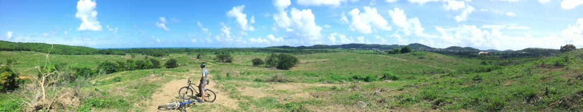 A panoramic view of a lush green landscape featuring rolling hills and a clear blue sky with fluffy white clouds. In the foreground, a person stands next to a mountain bike, looking out over the expansive scenery. The area appears to be open and uncrowded, showcasing the beauty of nature. Corredor Ecologico mountain bike trail.