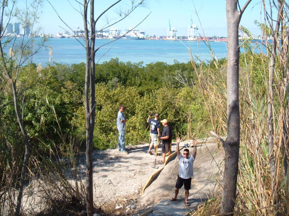Group of people standing on a rocky path surrounded by greenery, with a bay and shipping cranes visible in the background under a clear blue sky. One person is waving at the camera while others appear to be engaged in different activities. Virginia Key North Point mountain bike trail.