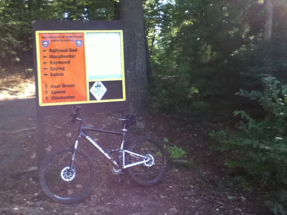 A mountain bike is leaning against a trail sign in a wooded area. The sign displays various trail names and directions, including Railroad Bed, Manchester, Raymond, and Bear Brook, in a vibrant orange and yellow format. Lush green trees and underbrush surround the scene, indicating a natural outdoor environment. Tower Hill Pond mountain bike trail.