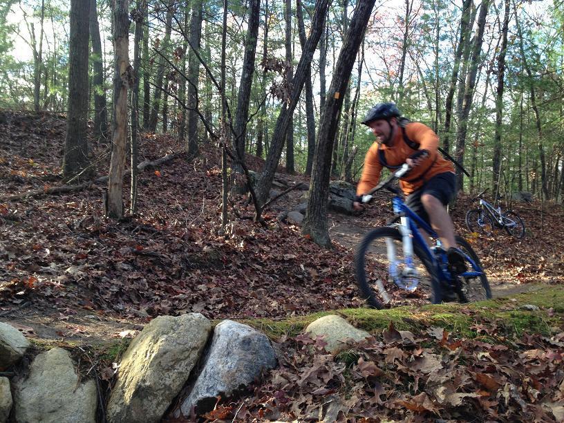 A person wearing an orange shirt and a helmet rides a mountain bike along a trail in a forest, navigating through fallen leaves and rocks. In the background, another bicycle is visible, surrounded by trees showcasing autumn foliage. Russell Mill mountain bike trail.