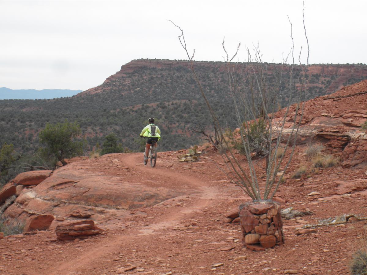 A cyclist riding on a dirt trail through a rugged, red rock landscape, surrounded by sparse vegetation and distant hills under a cloudy sky. Mescal Trail mountain bike trail.