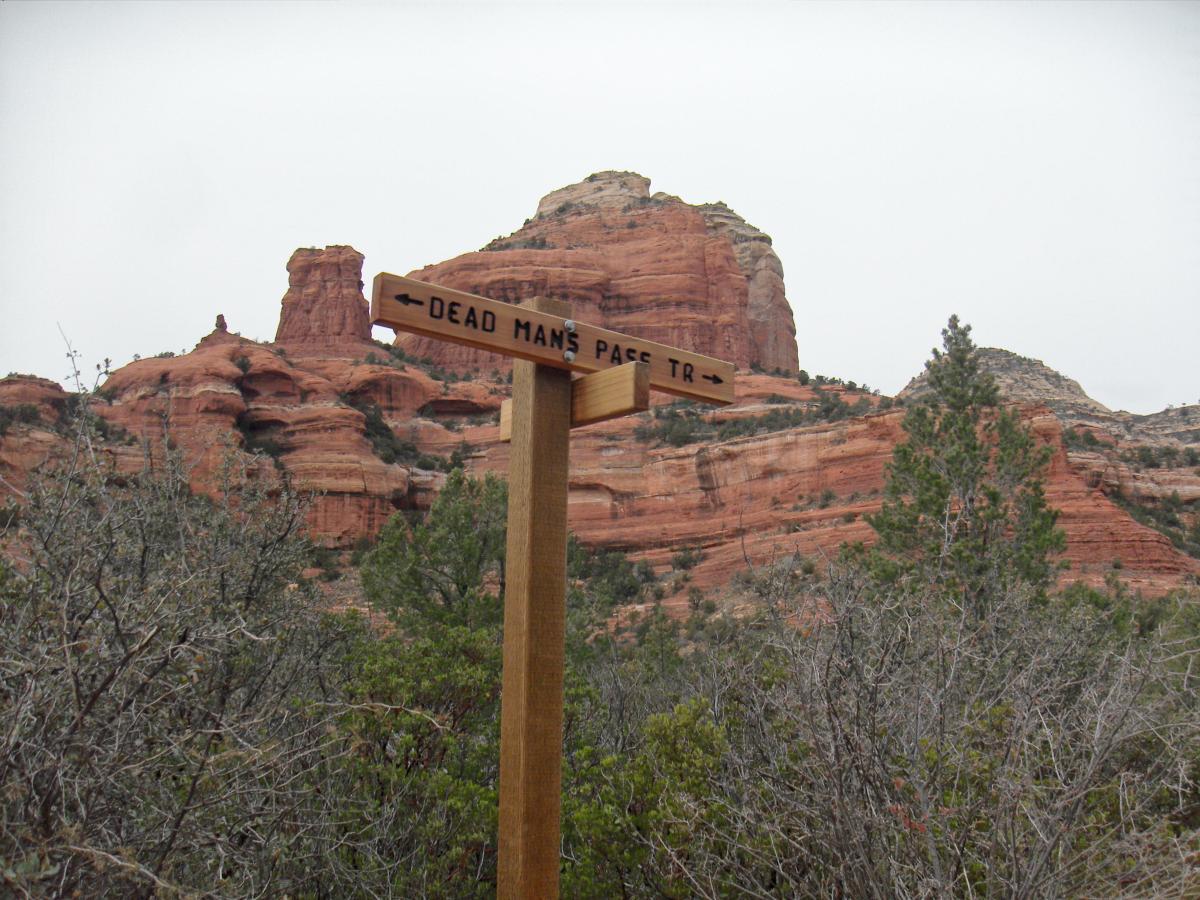 Wooden trail sign pointing towards "Dead Man's Pass Trail," surrounded by red rock formations and sparse greenery under an overcast sky. Mescal Trail mountain bike trail.