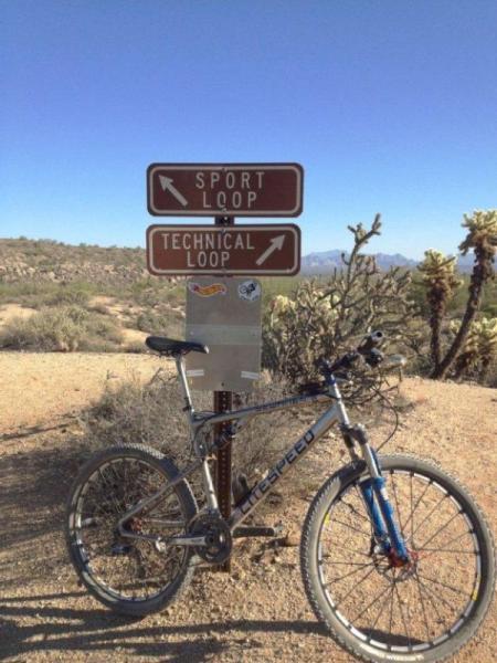 A mountain bike parked near a trail sign indicating directions to the "Sport Loop" and "Technical Loop" in a desert landscape with sparse vegetation and a clear blue sky. McDowell Mountain Park mountain bike trail.