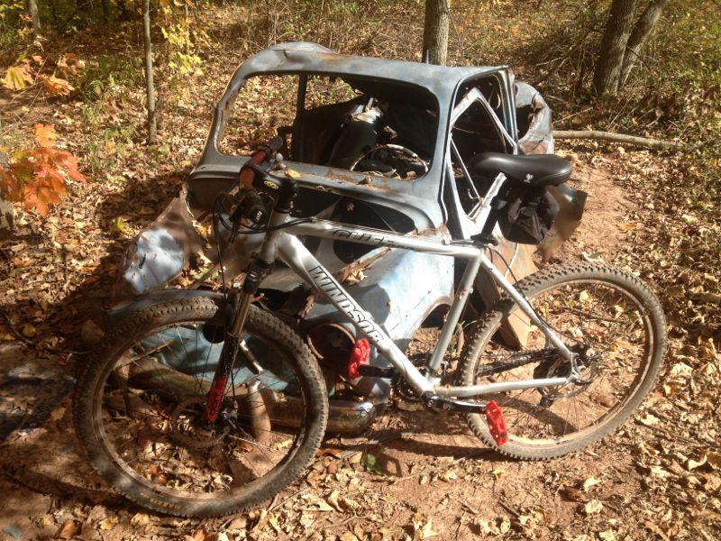A mountain bike parked beside a partially rusted and abandoned car in a wooded area, surrounded by fallen leaves and autumn foliage. Six Mile Run mountain bike trail.