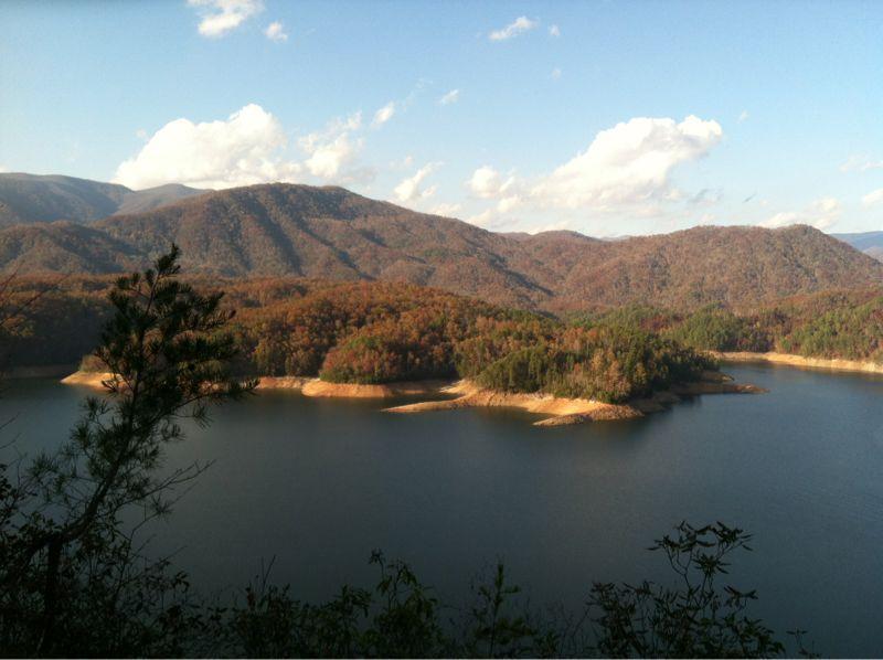 A scenic view of a tranquil lake surrounded by rolling hills and mountains, showcasing vibrant autumn foliage. The calm water reflects the blue sky and scattered clouds, while green trees and sandy shores create a peaceful natural landscape. Tsali Recreation Area mountain bike trail.