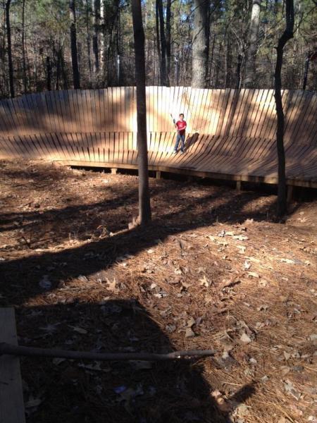A child stands on a wooden structure in a wooded area, surrounded by tall trees and pine needles on the ground. The structure features a curved design, resembling a half pipe, with a smooth wooden surface. The sunlight filters through the trees, casting shadows on the ground. Blankets Creek mountain bike trail.