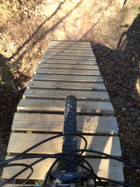A mountain bike is positioned at the top of a wooden ramp, with a view looking down the slope. The ground below is dirt and scattered with leaves, surrounded by trees. The bike's front wheel is clearly visible, showing the tread pattern. Sunlight filters through the trees, casting shadows on the ramp. Blankets Creek mountain bike trail.